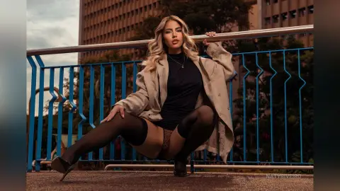 Stylish woman in a black bodysuit and beige blazer posing on a city balcony at sunset.