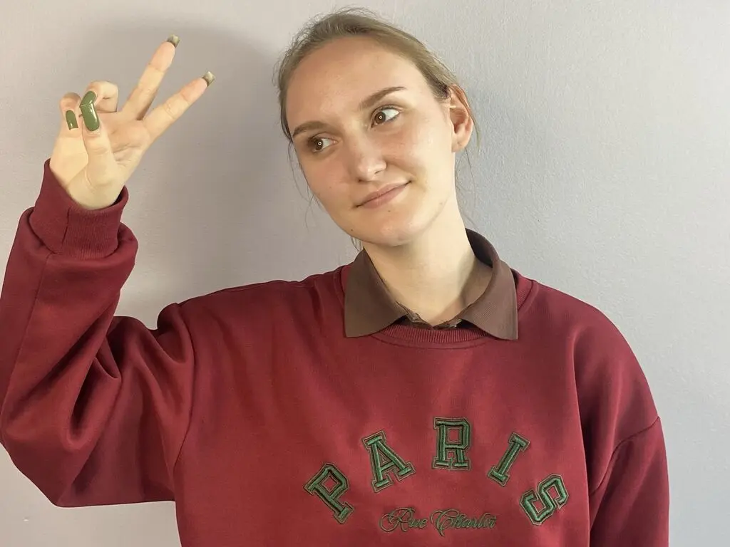 Smiling young woman in a burgundy Paris sweatshirt making a peace sign against a plain wall.