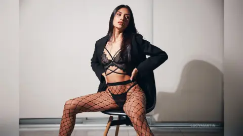 LucyMartin poses in black lace lingerie and a black blazer while wearing fishnet stockings, sitting on a chair against a plain wall.