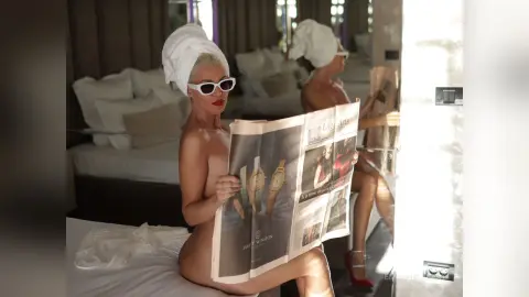 Blonde woman in a white towel and sunglasses reading a newspaper in a bright, modern bedroom.