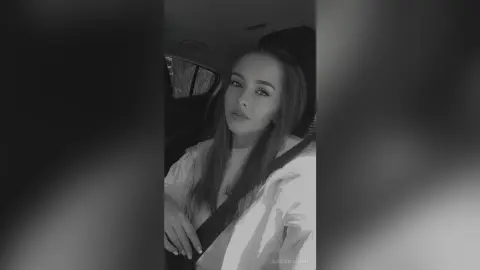 Black and white portrait of a young woman in a white top sitting inside a car with a moody expression.