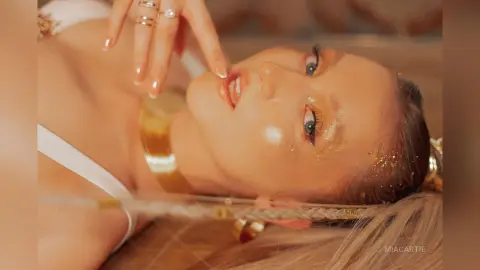 Glamorous woman with glitter makeup and gold jewelry lying down in a dreamy, ethereal setting.