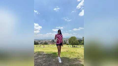 Stylish woman in a red patterned top and black shorts standing on a grassy hill under a blue sky.