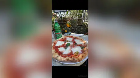 Freshly baked Margherita pizza with basil on a table next to a green soda bottle in a sunny outdoor cafe setting.