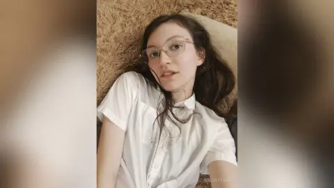 MillieStrife wearing glasses and a white button-up shirt while lying back on a tan pillow and bed.