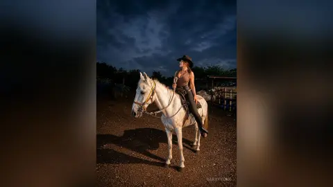 Cowgirl in a brown tank top and hat riding a white horse in a ranch setting under a moody twilight sky.
