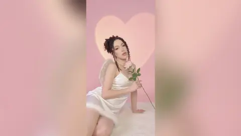 Dreamy portrait of a woman in a white satin dress holding a rose against a soft pink heart background.