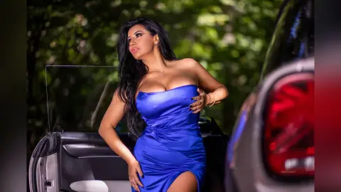 MissyJolie poses in a blue strapless dress next to a car in a wooded outdoor setting.