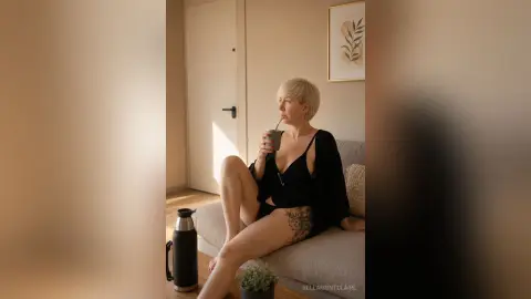 BellaMontclaire sits on a couch in a black top and shorts, sipping a drink from a cup.