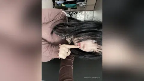 MarieJosePhilip poses with long black hair and a brown long-sleeve top, resting her chin on her hand against a dark background.