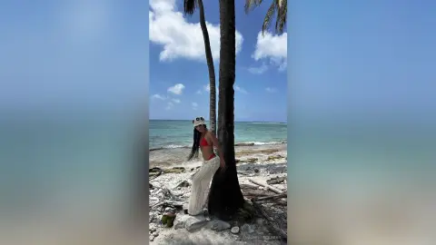 Woman in white crochet pants and red bikini top posing on a sunny tropical beach by palm trees.