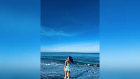 Young woman in a mint green swimsuit looking at the ocean under a clear blue sky at a sunny beach.