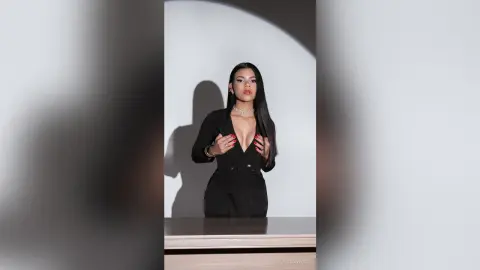 Elegant woman in a black plunging jumpsuit and pearl necklace posing against a minimalist grey studio backdrop.