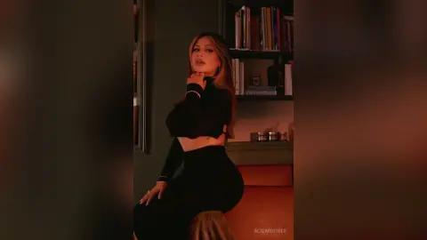Stylish woman in a black long-sleeve crop top posing elegantly in a moody, dimly lit room with bookshelves.