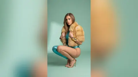 Glamorous woman in a teal bodysuit and tan faux fur coat posing against a mint green studio background.