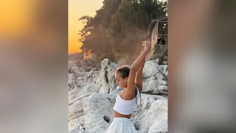 Young woman in white summer outfit stretching on rocky coast during a golden sunset.