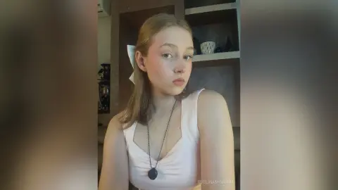 EvelinaMoorina poses in a white tank top and necklace, looking at the camera with her blonde hair tied back with a white bow.
