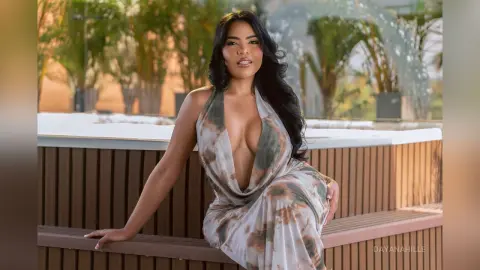 DayanaHille poses in a low-cut, tie-dye patterned dress that shows deep cleavage, sitting on a wooden bench outdoors.