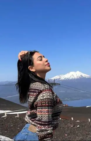Young woman in patterned bohemian sweater relaxing under blue sky near snow-capped mountains.