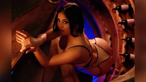 Glamorous woman in a beige bodysuit posing in a moody, dimly lit wine cellar surrounded by wooden barrels.