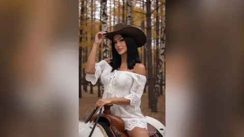 Woman in white lace boho dress and brown cowboy hat sitting in a dreamy autumn forest setting.