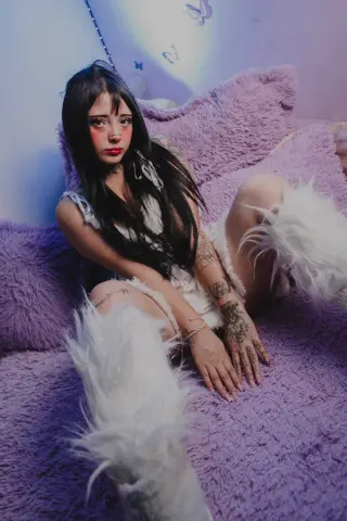 Young woman with long dark hair in a white fuzzy outfit sitting on a purple shag rug in a dreamy bedroom.