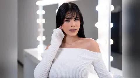 Beautiful brunette woman in a white off-the-shoulder sweater posing in a bright vanity studio setting.