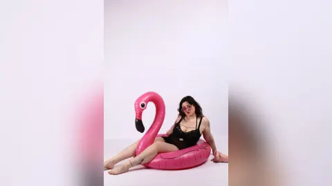 KiraKovall wearing a black lace bodysuit while sitting inside a large pink inflatable flamingo float against a white background.