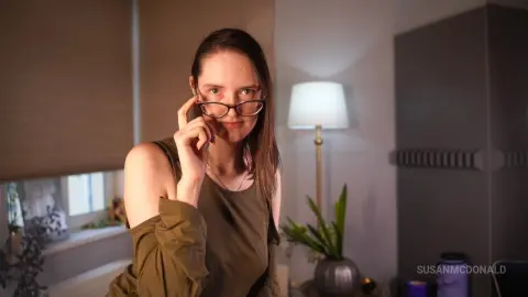 Young woman in an olive green top adjusting glasses in a cozy, dimly lit room.
