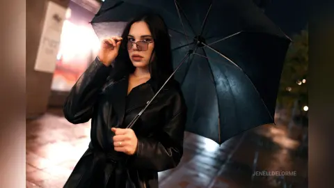 JenellDelorme poses outdoors at night wearing a black leather jacket and sunglasses while holding a large black umbrella.