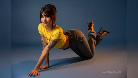 Young woman in yellow t-shirt and dark jeans posing playfully on a blue studio background.