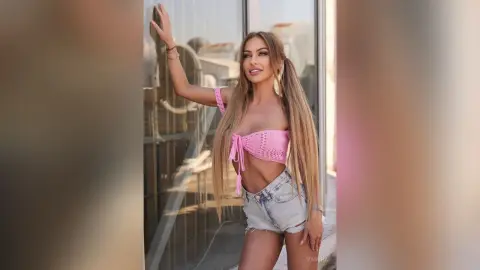 Beautiful woman in pink crochet crop top and denim shorts posing by a glass door in warm sunlight.