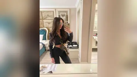 ArianaKroft poses in a black long-sleeved crop top and black leggings, holding a phone for a mirror selfie in a bedroom.