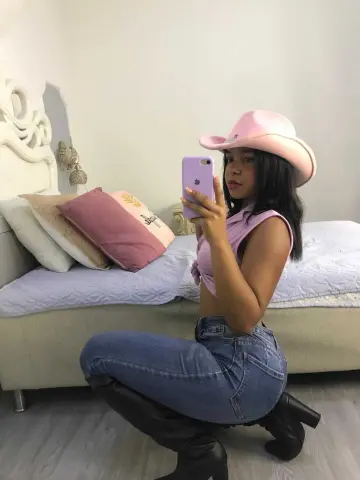 Young woman in pink crop top, blue jeans, and cowboy hat posing in a bedroom.