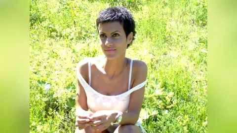 Smiling woman in a white tank top sitting in a sunny green meadow field.