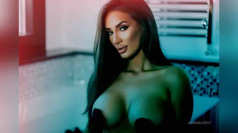 AnnaKarev poses with long dark hair and heavy makeup, showing off her large breasts in a dimly lit bathroom.