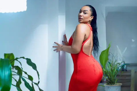 Curvy woman in a red backless dress posing in a bright room with green houseplants.