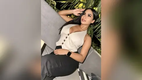 MiaKorz poses on a grey outdoor sofa wearing a low-cut white sleeveless top and black pants, looking at the camera with long dark hair.