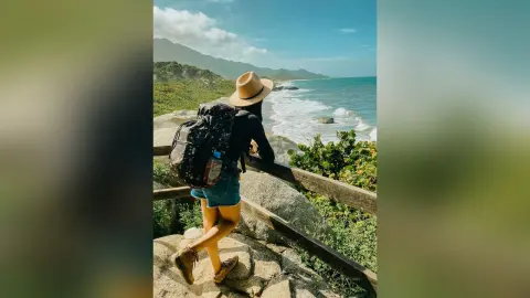 Adventurous woman with a black backpack and sun hat hiking along rocky coastal cliffs overlooking the ocean.