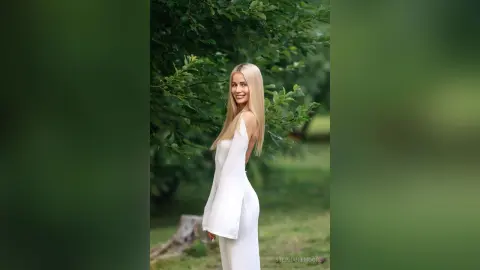 Smiling blonde woman in a white elegant dress standing in a lush green garden setting.