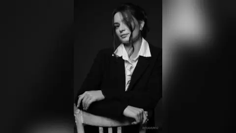 Elegant woman in a black blazer and white shirt posing in a moody, dark studio setting.