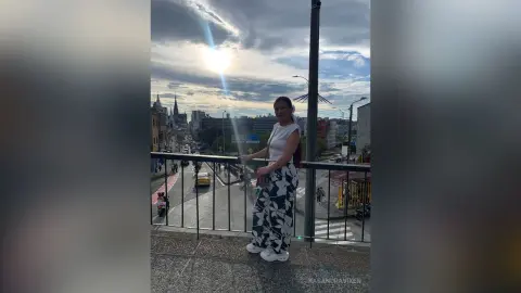 Young woman in white floral trousers standing on a city balcony under a dramatic sunset sky.