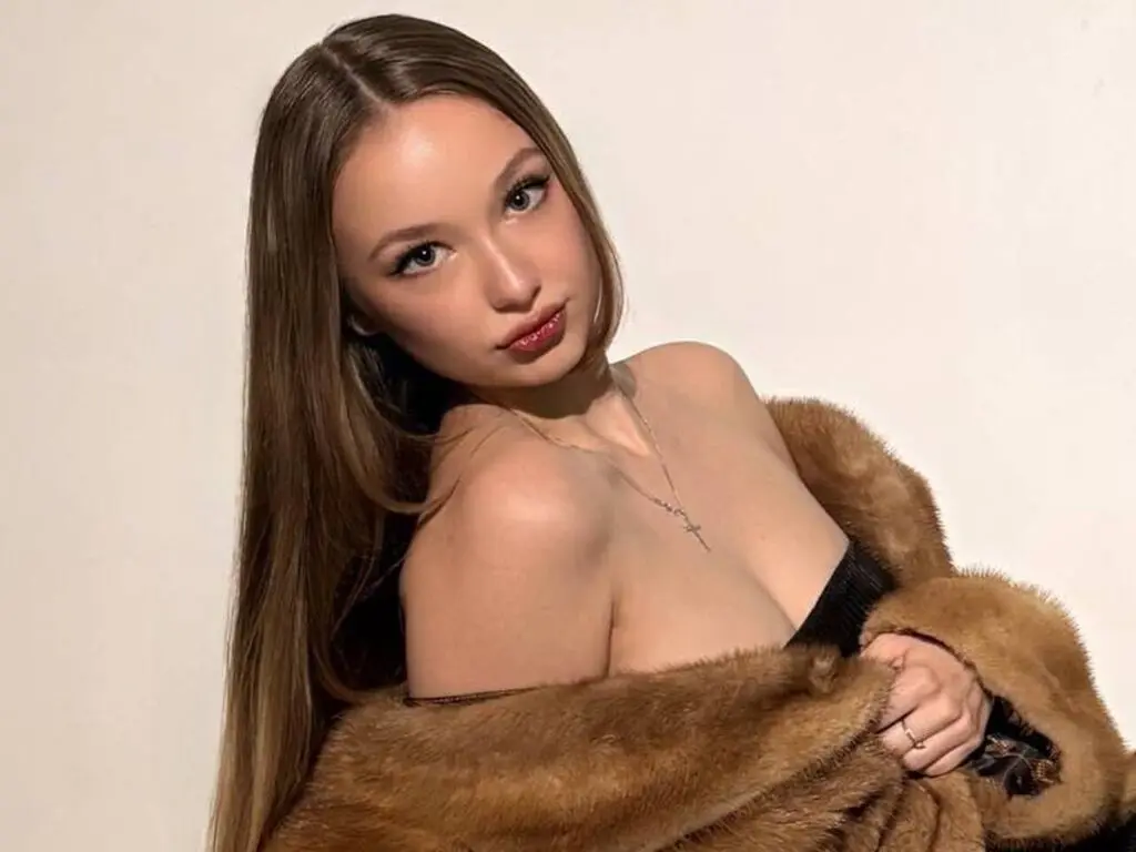 Young woman with long brown hair wearing a brown faux fur coat and necklace against a white background.