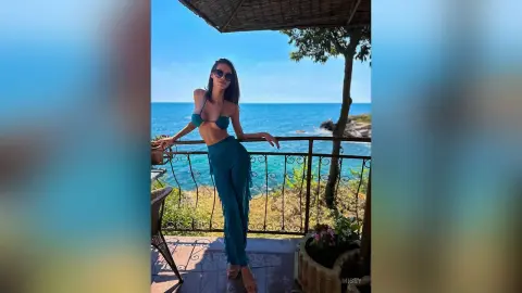 Stylish woman in a teal bikini top and matching trousers posing on a sunny seaside balcony overlooking the ocean.