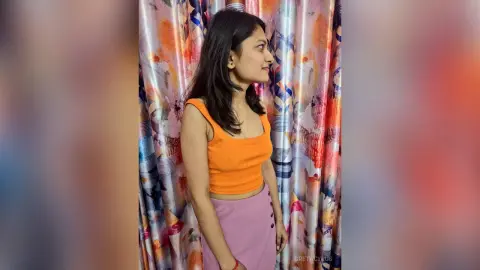 Young woman in an orange ribbed tank top and pink skirt posing against colorful patterned curtains.
