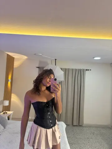 Young woman in a black corset and beige skirt taking a selfie in a cozy, warmly lit bedroom.