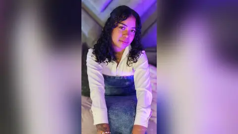 NaomiVelour poses in a white long-sleeve shirt and denim skirt, showing off her dark curly hair and tattoos.