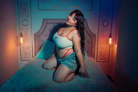 Young woman in white crop top and denim shorts sitting on a bed in cozy, moody teal lighting.