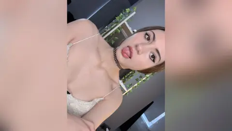 AriaLush wearing a white lace bra, sticking her tongue out while looking at the camera in a room with green leafy plants behind her.
