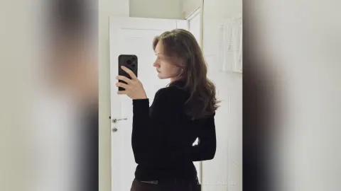 GwendolynBucey poses for a mirror selfie wearing a black long-sleeved shirt, showing her long brown hair and profile against a white door and wall.